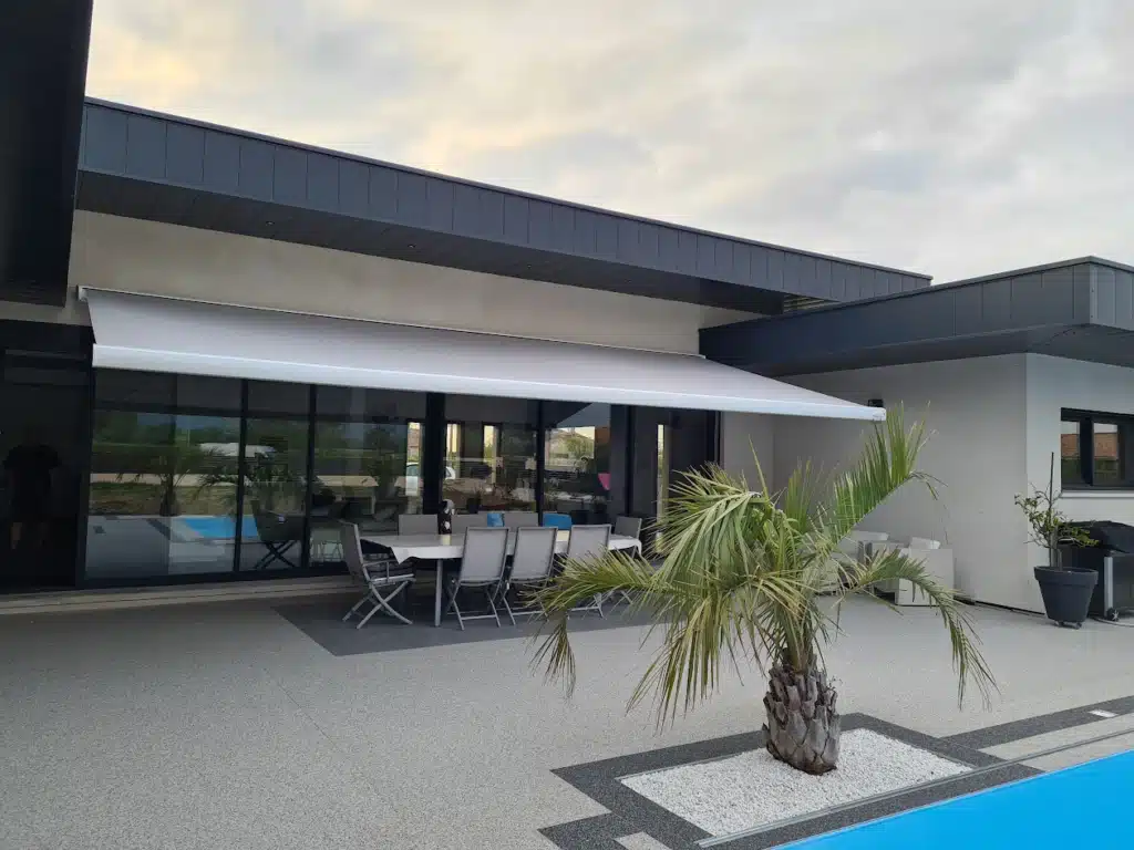 Terrasse moderne avec store banne rétractable gris clair, mobilier de jardin et palmier près d'une piscine au crépuscule.